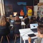 : Grupa dzieci i młodzieży uczestniczy w warsztatach prowadzonych przez Fundację GraTy, słuchając prelegentów i korzystając z komputerów.