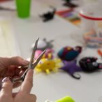 Ręce osoby trzymającej nożyczki i przygotowującej materiały do rękodzieła. Na stole widoczne są kolorowe ozdoby oraz narzędzia potrzebne do pracy.