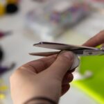 Zbliżenie na rękę osoby wycinającej kawałek materiału za pomocą nożyczek, przygotowując element do pracy rękodzielniczej