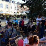 Ludzie siedzący przy stolikach na rynku, grający w gry planszowe i uczestniczący w warsztatach partycypacyjnych w ramach kawiarni obywatelskiej.