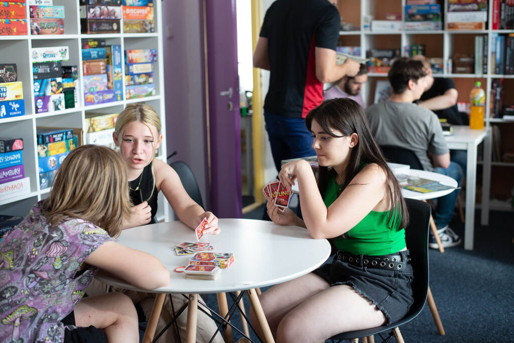 Trzy młode osoby siedzą przy okrągłym stole, grając w karty, w tle widoczne są regały pełne gier planszowych.