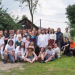 Grupa ludzi w różnym wieku pozuje do zdjęcia na zewnątrz, na tle zielonego ogrodu i drewnianego budynku.