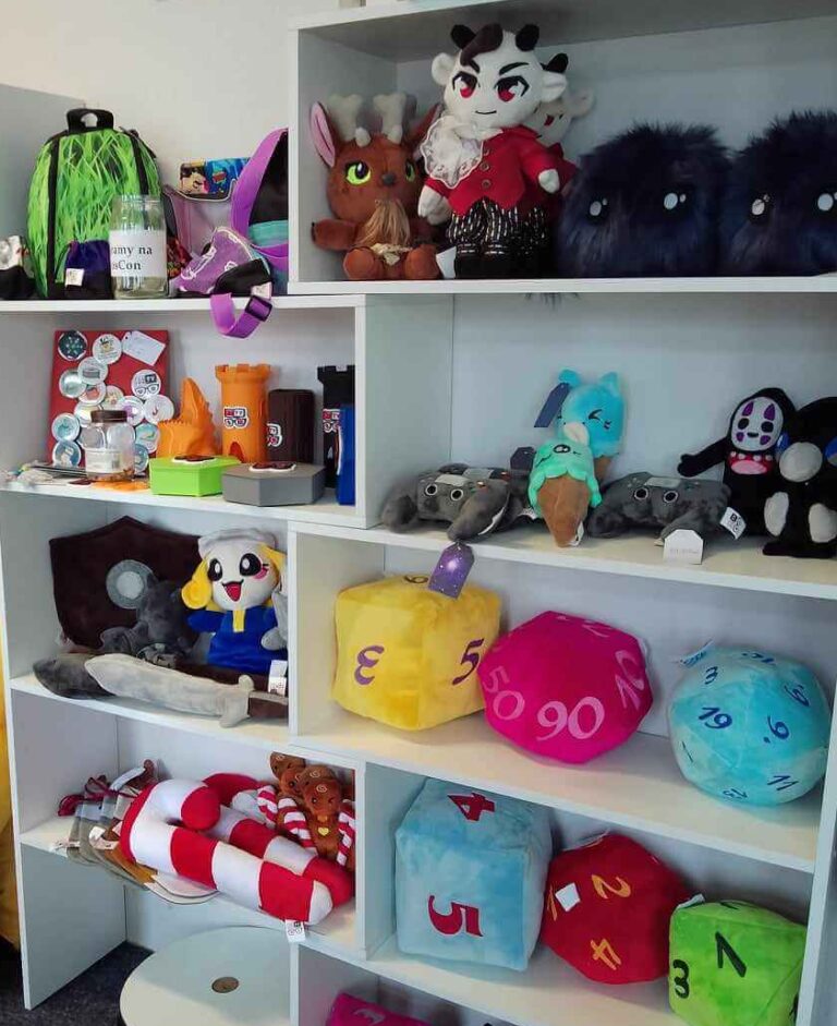 Półka z różnorodnymi produktami stworzonymi przez Fundację GraTy. Na półkach znajdują się pluszowe kostki do gry, maskotki, akcesoria i gadżety związane z grami.