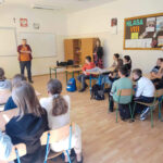 Mężczyzna w pomarańczowej koszuli prowadzi spotkanie w klasie szkolnej. Uczniowie siedzą przy biurkach i słuchają z zainteresowaniem. Na tablicy korkowej w tle widać napis "KLASA VIII".