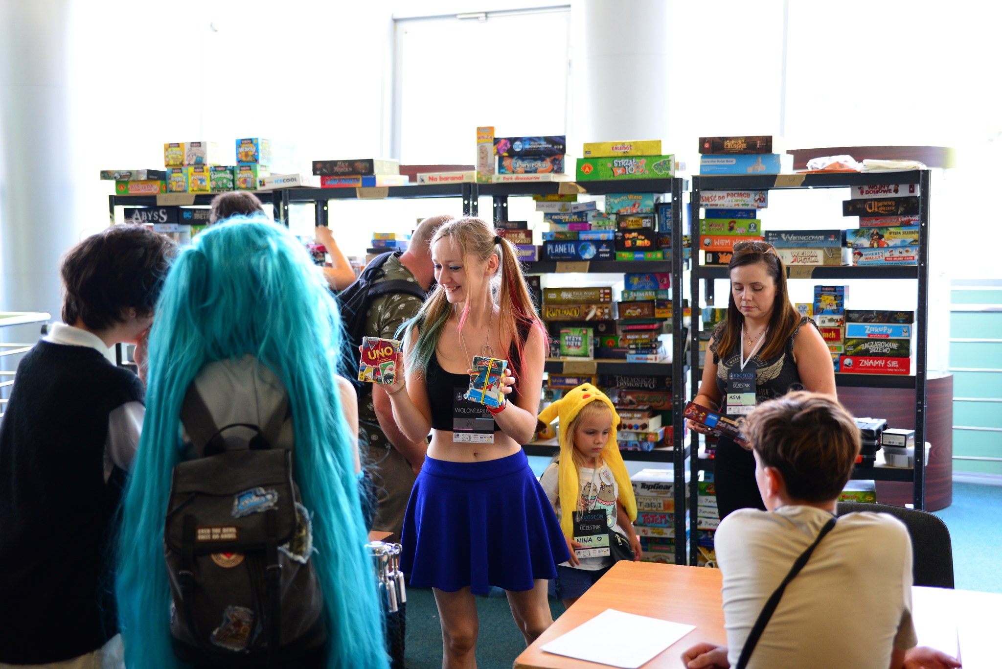 Na zdjęciu widać radosną, uśmiechniętą kobietę z jasnymi kucykami, która w otoczeniu grupy entuzjastycznych ludzi poleca grę do wypożyczenia. W tle zauważyć można regały zapełnione różnorodnymi planszówkami, które czekają na chętnych do wypożyczenia. Atmosfera jest pełna entuzjazmu i gotowości do spędzenia czasu z przyjaciółmi przy planszówkach