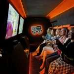 Na zdjęciu widać grupę dorosłych osób bawiących się we wnętrzu GRAtoWOZu. Siedzą wygodnie na kanapach, trzymając w rękach kontrolery do konsoli. Ich twarze są skoncentrowane, a jednocześnie pełne radości z rozgrywki. Pomieszczenie jest oświetlone pomarańczowymi światłami, co nadaje mu ciepły i przyjazny klimat. W tle widoczny jest telewizor, na którym wyświetlają się emocjonujące sceny z gier. Cała scena emanuje energią i dobrą atmosferą.