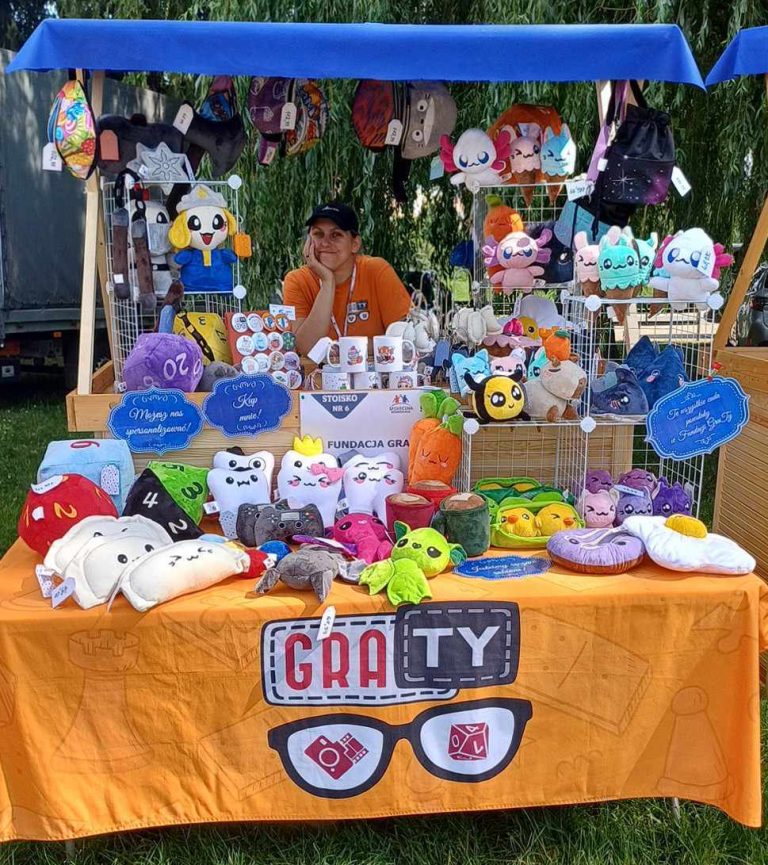 Na zdjęciu znajduje się stoisko Fundacji GraTy, na którym prezentowane są różnorodne pluszowe zabawki i akcesoria. Na stole widoczne są maskotki w kształcie zwierząt, geometryczne pluszowe kostki oraz inne kolorowe przedmioty, które przyciągają uwagę odwiedzających.