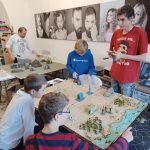 Kilku młodych chłopców uczestniczy w rozgrywce gry bitewnej na dużej makiecie. Starszy chłopak w czerwonej koszulce trzyma notes i zapisuje notatki, a w tle dorosły mężczyzna przygotowuje dodatkowe elementy gry.