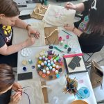 Trzy osoby ozdabiające materiałowe torby farbami, używając pędzli i różnych kolorów farb, siedzące wokół stołu pełnego przyborów plastycznych.