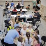 Grupa dzieci i dorosłych uczestniczących w warsztatach gier planszowych na świeżym powietrzu, przy stołach ustawionych na dziedzińcu.