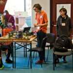 Trzech młodych uczestników stoi i siedzi przy stole z rozłożonymi figurkami i elementami gry bitewnej. W tle widoczny jest baner "Warhammer Age of Sigmar" oraz otwarte drzwi.