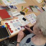 : Starsza kobieta siedzi przy stole, zajmując się pracami plastycznymi. Przed nią leżą różnorodne kolorowe materiały, takie jak kredki, pisaki i ozdobne papiery, które wykorzystuje do tworzenia artystycznych kompozycji. W tle widać dodatkowe materiały artystyczne, rozłożone na stole.