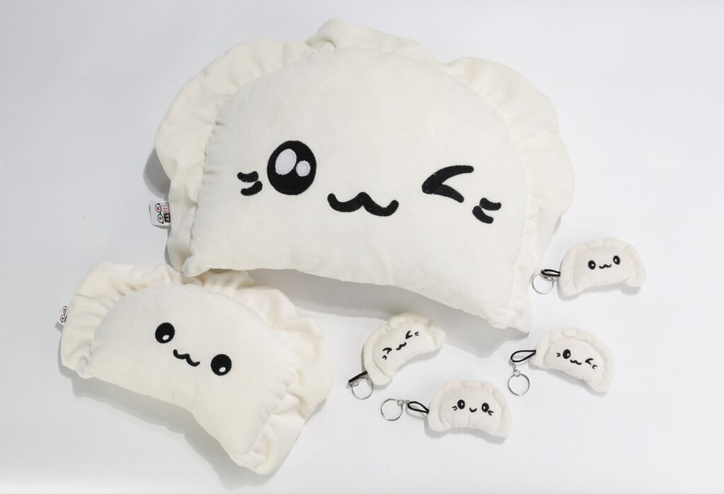 Pluszowe pierogi: dwie duże, kremowe poduszki w kształcie pierogów z czarnymi, haftowanymi twarzami, jedna z otwartymi oczami, druga z jednym okiem zamkniętym, oraz cztery małe pluszowe pierogi z czarnymi, haftowanymi twarzami, każdy z kółkiem do kluczy, leżące na jasnoszarym tle.