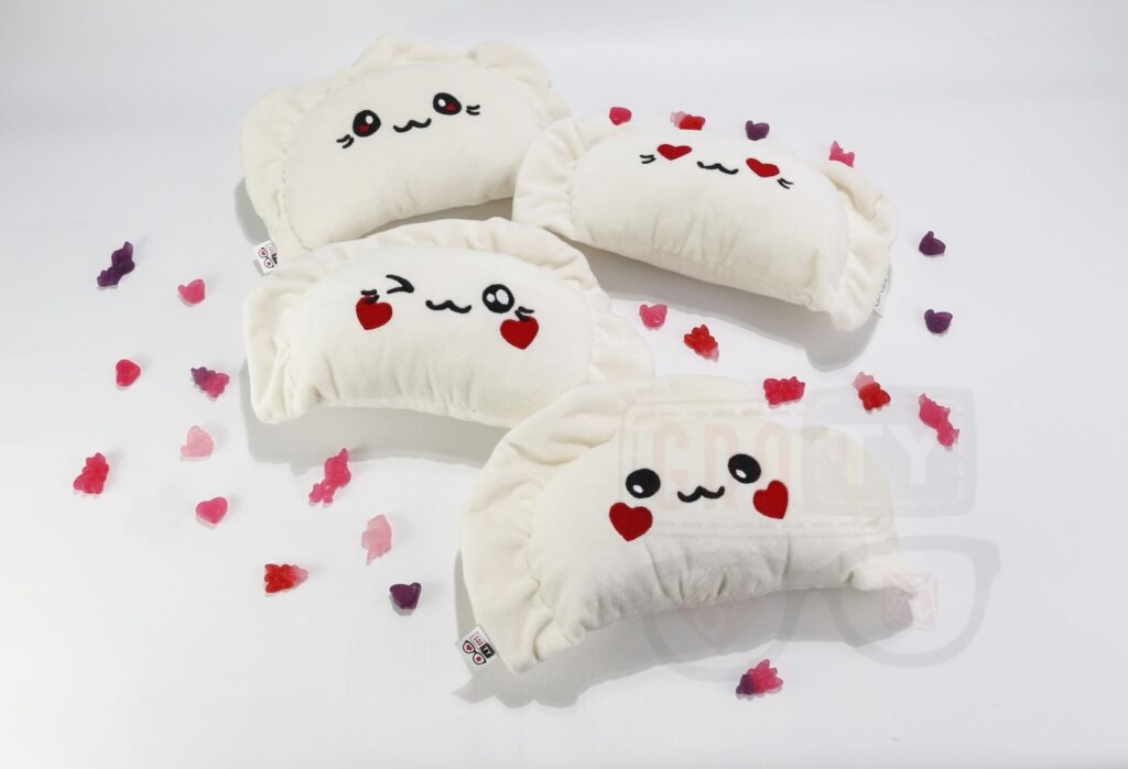 Cztery pluszowe pierogi z walentynkowymi akcentami: kremowe pierogi z czarnymi, haftowanymi twarzami i czerwonymi sercami zamiast oczu, otoczone czerwonymi i różowymi, pluszowymi serduszkami, leżące na jasnoszarym tle.