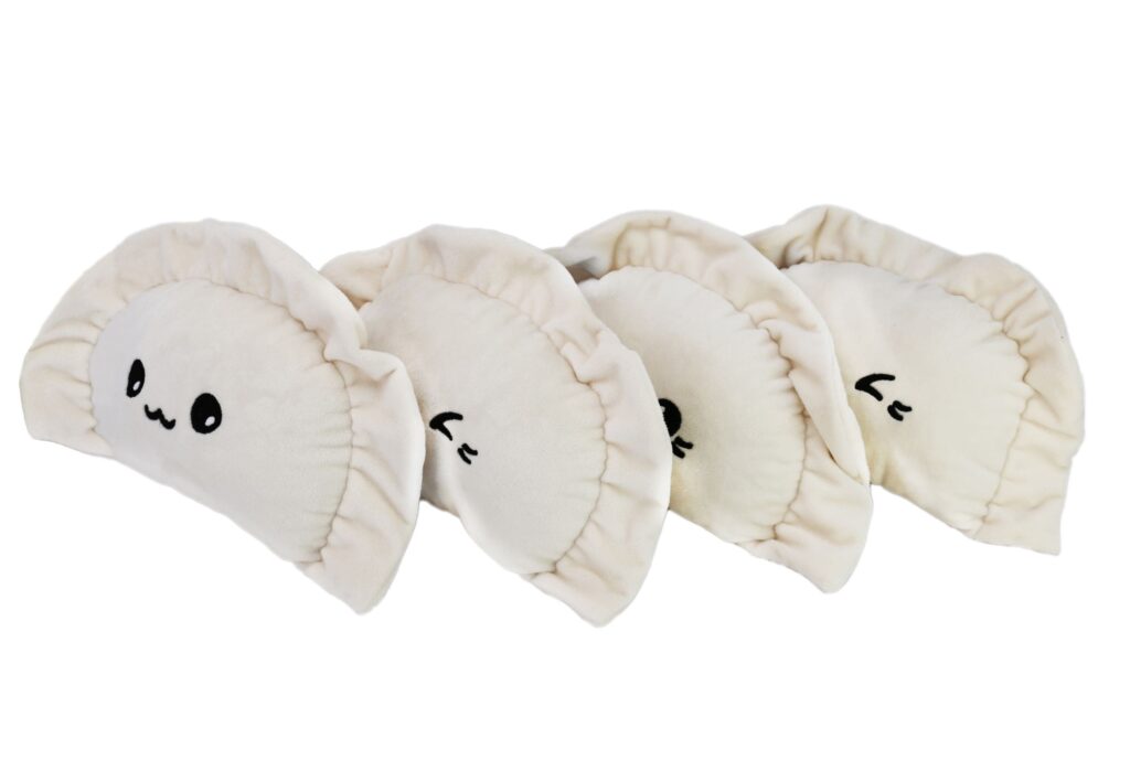 Cztery pluszowe pierogi ułożone w rzędzie: kremowe pierogi z czarnymi, haftowanymi twarzami, każdy z innym wyrazem, leżące na jasnoszarym tle.