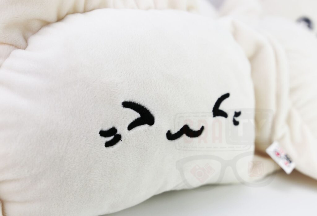 Zbliżenie na pluszowego pieroga: kremowy pieróg z czarną, haftowaną twarzą z jednym okiem zamkniętym i uśmiechniętymi ustami, widoczne włókna pluszu i metka, leżący na białym tle.