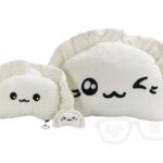 Pluszowe pierogi: dwa duże, kremowe pierogi z czarnymi, haftowanymi twarzami, jeden z otwartymi oczami, drugi z jednym okiem zamkniętym, oraz mały pluszowy pieróg z kółkiem do kluczy, leżące na jasnoszarym tle.