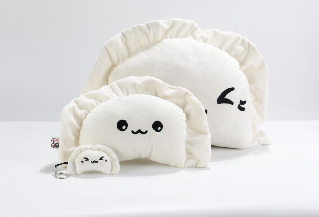 Pluszowe pierogi: dwa duże, kremowe pierogi z czarnymi, haftowanymi twarzami, jeden z otwartymi oczami, drugi z jednym okiem zamkniętym, oraz mały pluszowy pieróg z kółkiem do kluczy, leżące na jasnoszarym tle.