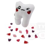 Pluszowy ząb z haftowanymi czerwonymi sercami zamiast oczu i czarnym uśmiechem, z którego spadają kolorowe, pluszowe serduszka, na białym tle.