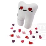 Pluszowy ząb z haftowanymi czerwonymi sercami zamiast oczu i czarnym uśmiechem, z którego spadają kolorowe, pluszowe serduszka, leżący na białym tle.