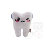 Pluszowy ząb z niebieskimi skrzydełkami, haftowanymi czerwonymi sercami zamiast oczu i czarnym uśmiechem, leżący na białym tle.