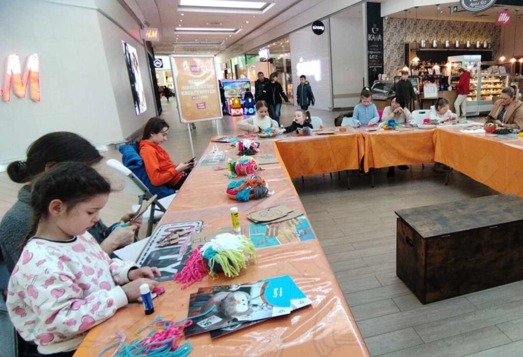 Grupa dzieci i dorosłych siedzi przy stołach ustawionych w literę U, w przestrzeni centrum handlowego. Na stołach znajdują się kolorowe włóczki, kleje i materiały plastyczne. Uczestnicy skupiają się na tworzeniu prac rękodzielniczych.