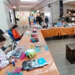 Grupa dzieci i dorosłych siedzi przy stołach ustawionych w literę U, w przestrzeni centrum handlowego. Na stołach znajdują się kolorowe włóczki, kleje i materiały plastyczne. Uczestnicy skupiają się na tworzeniu prac rękodzielniczych.