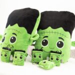 Na zdjęciu widoczne są pluszowe maskotki przedstawiające zielone postacie inspirowane Frankensteinem. Dwie duże poduszki mają czarne „włosy”, wielkie oczy, uśmiechnięte buzie, zielone uszy i przyszyte po bokach szare „śruby”. Przed nimi stoi sześć mniejszych maskotek – breloczków w tym samym stylu.