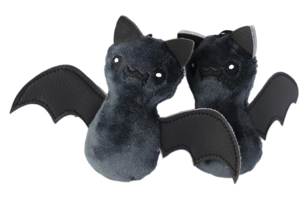 Dwa pluszowe nietoperze w ciemnoszarym kolorze, z dużymi, czarnymi skrzydłami wykonanymi z materiału przypominającego skórę. Mają czarne uszy, jasne oczy i uśmiechnięte buźki.