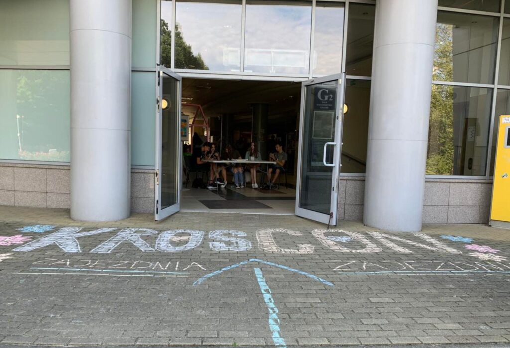 Wejście do budynku z otwartymi drzwiami, przed którymi na chodniku kolorową kredą napisano „KrosCon” oraz „zajezdnia” z dużą strzałką kierującą do środka. W tle widać uczestników siedzących przy stole wewnątrz budynku.