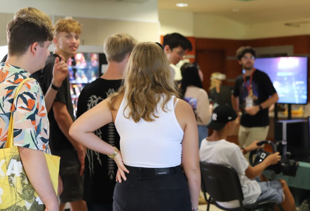 Grupa młodych osób zgromadzona wokół stanowisk z monitorami i sprzętem do gier. W tle osoba siedzi przy symulatorze jazdy, obok stoi prowadzący w czarnej koszulce z identyfikatorem.