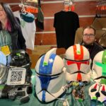 Dwójka wystawców stoi za stołem pełnym hełmów inspirowanych Gwiezdnymi Wojnami. Na stole leżą kolorowe repliki hełmów klonów, figurki, gadżety i tabliczka z kodem QR.