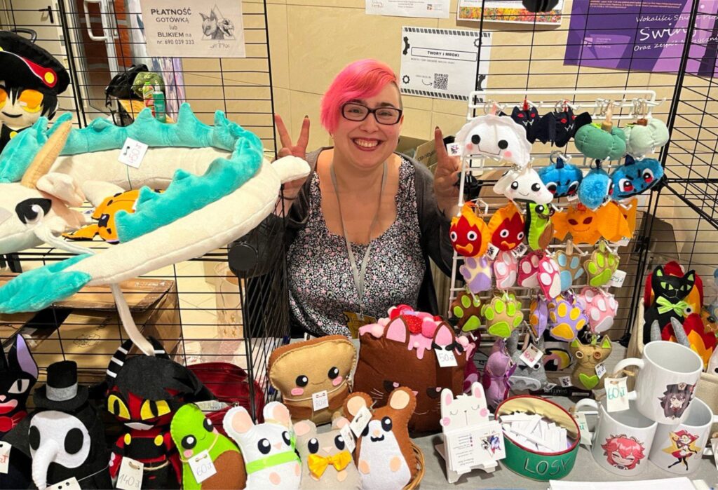 Uśmiechnięta kobieta z różowymi włosami stoi za stoiskiem z ręcznie robionymi pluszakami i gadżetami. Na metalowych stojakach zawieszone są kolorowe maskotki przypominające smoki, koty i fantastyczne stworzenia.