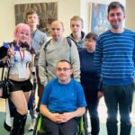 Grupa siedmiu osób pozuje wspólnie do zdjęcia w jasnym pomieszczeniu. Wśród nich osoba w cosplayu z różowymi włosami i strojem fantasy oraz osoba na wózku inwalidzkim. W tle wiszą obrazy i widać drzwi.