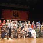 Grupa kilkudziesięciu osób w różnorodnych kostiumach cosplay pozuje na scenie. Wśród przebrań znajdują się postacie z anime, fantasy, science-fiction i militariów. W tle zawieszony jest baner z czarno-czerwonym symbolem.