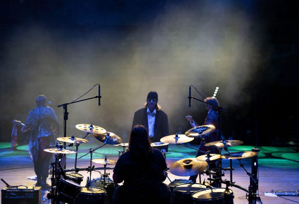 Widok ze sceny na występ muzyczny – z przodu perkusista, a dalej trzech muzyków stojących w świetle reflektorów i otoczeniu dymu scenicznego. W tle widać kolorowe oświetlenie i zarys publiczności.