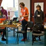 Trzech młodych chłopaków stoi i siedzi wokół stołu z planszą do gry bitewnej Warhammer. Na stole ustawione są modele budynków i figurek. W tle widoczny jest baner Warhammer Age of Sigmar.