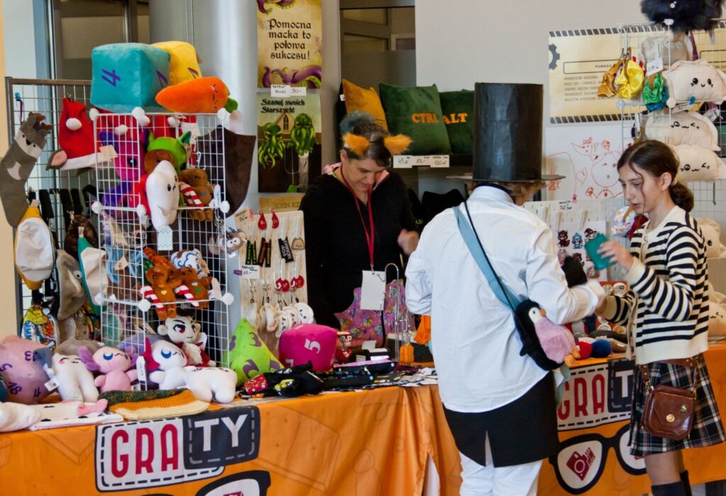 Stoisko wystawiennicze Fundacji GraTy z ręcznie wykonanymi maskotkami, torbami i ozdobami. Przed stołem dwie osoby przeglądają produkty – jedna z nich w cylindrze, druga w spódniczce w kratę. W tle osoba z opaską z uszami obsługuje stoisko.