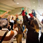 Grupa osób w kolorowych strojach cosplay stoi w kręgu z uniesionymi rękami, trzymając pluszowe maskotki. W środku postać w masce gazowej. Jedna osoba nagrywa scenę telefonem.