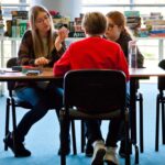 Trzy osoby siedzą przy stole i grają w grę planszową. W tle widać regał pełen pudełek z grami. Jedna z uczestniczek tłumaczy zasady, pokazując coś w dłoni.