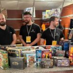 Trzech mężczyzn w czarnych koszulkach i identyfikatorach stoi za ladą wypełnioną grami planszowymi. Przed nimi ustawione są pudełka gier takich jak „Splendor”, „Tajniacy”, „Root” i inne.