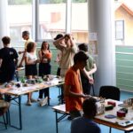 Grupa osób bierze udział w rozgrywkach bitewnych przy kilku stołach. Na stołach znajdują się makiety terenów i figurki. W tle widać ludzi rozmawiających i obserwujących, a całość odbywa się w jasnej sali z dużymi oknami.