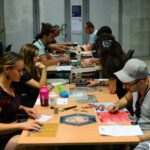 Kilkunastoosobowa grupa uczestników siedzi przy długich stołach i gra w planszówki. Na stole widać plansze, pionki, kostki i instrukcje do gier. W tle widać więcej osób grających w kolejnych rzędach.