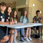 Grupa młodzieży siedzi przy stole rejestracyjnym w przestrzeni wewnętrznej budynku. Na stole leżą identyfikatory, długopisy, kartki i płyn do dezynfekcji. Z boku podchodzi uczestniczka wydarzenia w ciemnej koszulce i spodenkach moro.