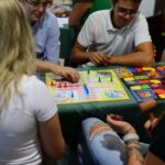 Cztery osoby siedzą przy stole i grają w kolorową planszową grę towarzyską. Na stole widać planszę z pionkami i kostkami, a obok inne gry. Uczestnicy są uśmiechnięci i zaangażowani w rozgrywkę.