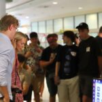 Grupa osób stoi w holu budynku, patrząc w stronę stanowiska komputerowego. Na pierwszym planie widać ekran monitora, a w tle – uczestników wydarzenia w różnorodnych strojach, m.in. w koszulkach z grafikami i opaskach.