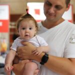 Mężczyzna w białej koszulce trzyma na rękach małe dziecko w jasnym body. Dziecko ma jasne włosy związane w mały kucyk na czubku głowy i patrzy prosto w obiektyw.