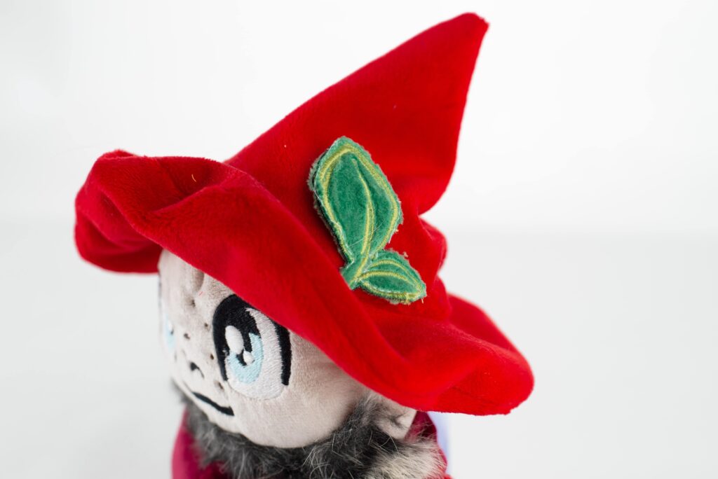 Zbliżenie na głowę pluszowej maskotki Krośnięcia – widoczny czerwony, spiczasty kapelusz z naszytym zielonym listkiem oraz fragment twarzy z dużym okiem.